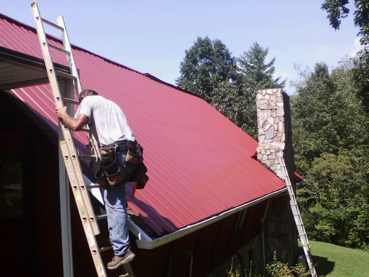 Roofer on a ladder beside a red metal roof and stone chimney — Soffit & Fascia Repair by Roofing Brothers in Harrison City